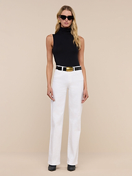 A woman wearing sunglasses, the Ceci Sleeveless Turtleneck in black, white pants with a wide belt, and black shoes stands against a plain white background.