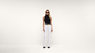 A woman wearing sunglasses, a sleeveless black top, white high-waisted pants, a wide belt, and black shoes stands against a plain white background.