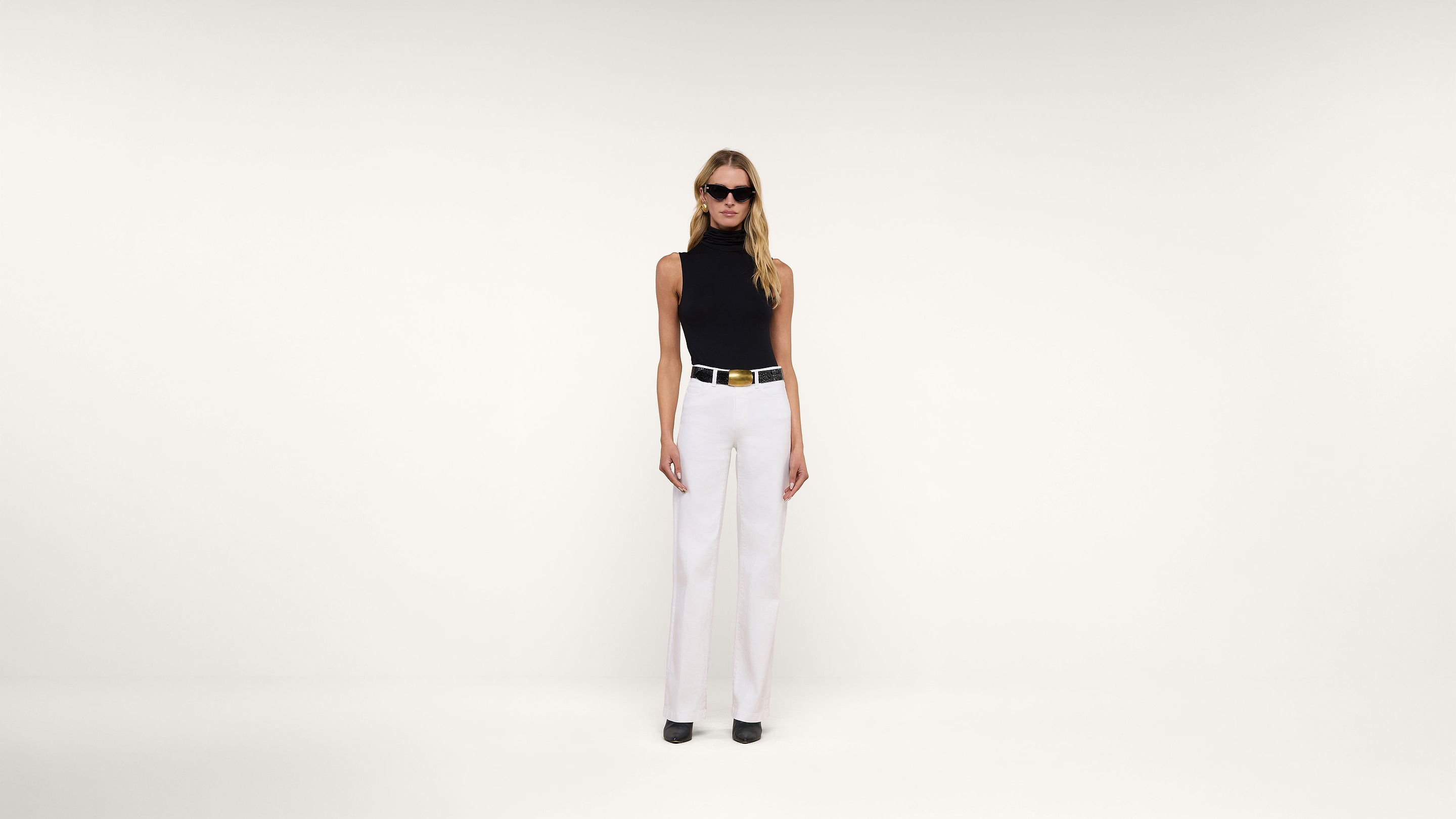 A woman wearing sunglasses, a sleeveless black top, white high-waisted pants, a wide belt, and black shoes stands against a plain white background.