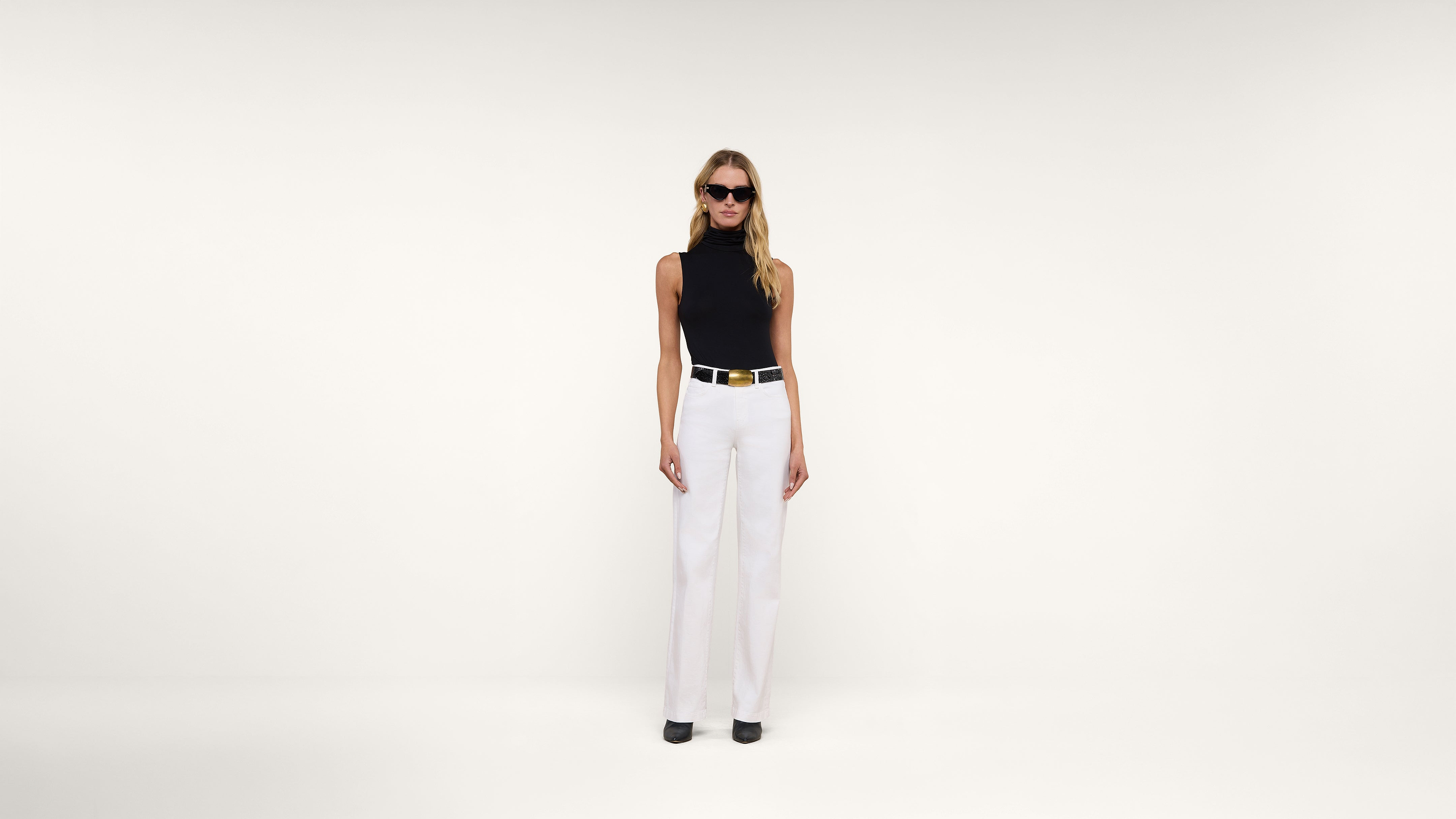 A woman wearing sunglasses, a sleeveless black top, white high-waisted pants, a wide belt, and black shoes stands against a plain white background.