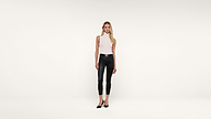 A woman stands against a plain white background, wearing a sleeveless white top, black leather pants, and black heels.