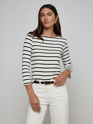 A woman with long brown hair wears the Lucille Striped Boatneck Top, styled with white pants, a black belt, and gold hoop earrings, standing against a plain light background.
