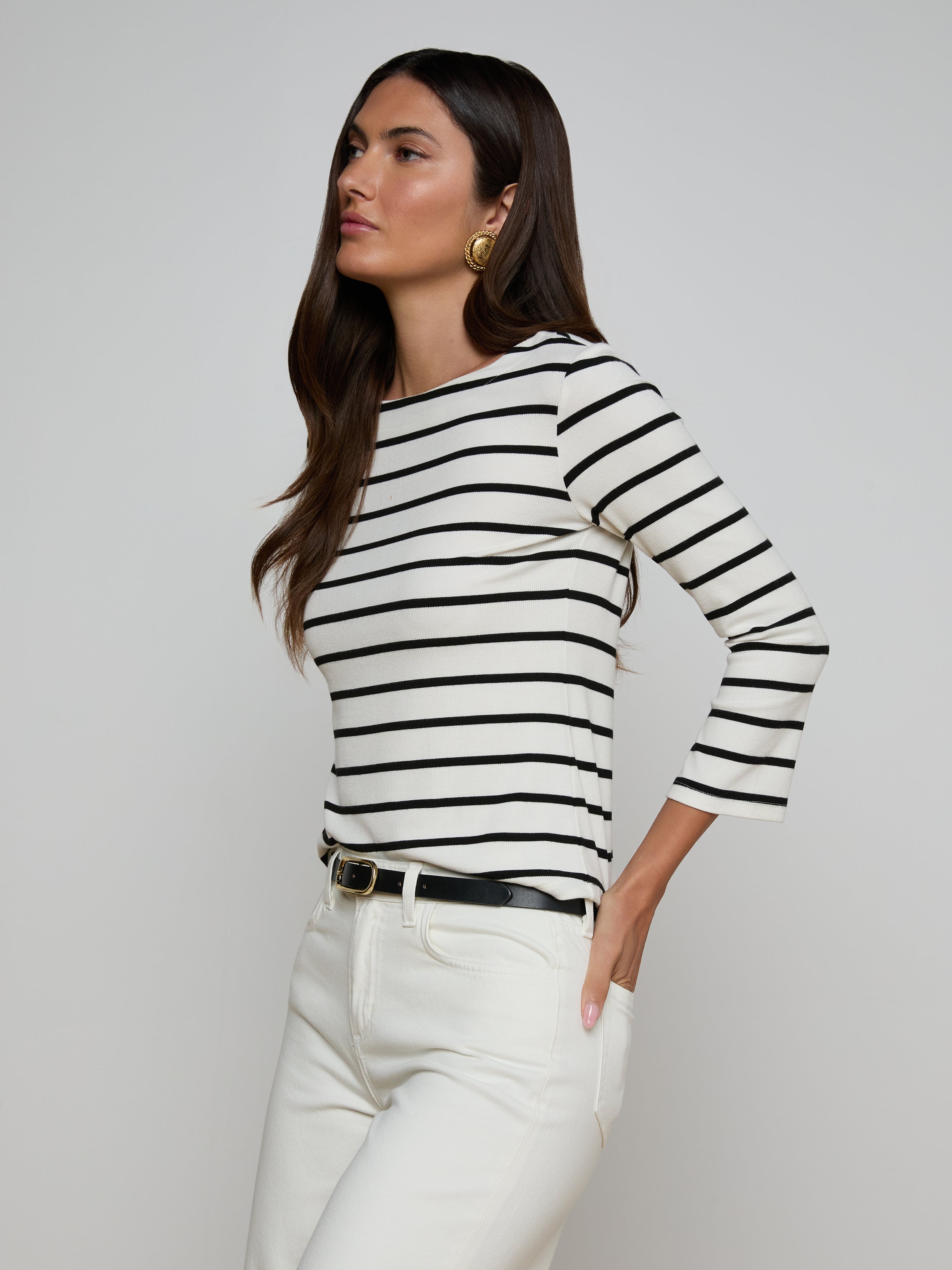Wearing the Lucille Striped Boatneck Top—a black and white, long-sleeve shirt featuring a boat neckline—paired with white pants and a black belt, the woman stands with one hand in her pocket against a plain background. The timeless stripes of the top add classic appeal to her look.
