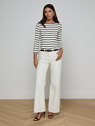 A woman stands against a plain background wearing the Lucille Striped Boatneck Top, paired with white wide-leg pants, a black belt, and black high-heeled sandals. With one hand in her pocket and a neutral expression, she captures the effortlessly chic appeal of timeless stripes.