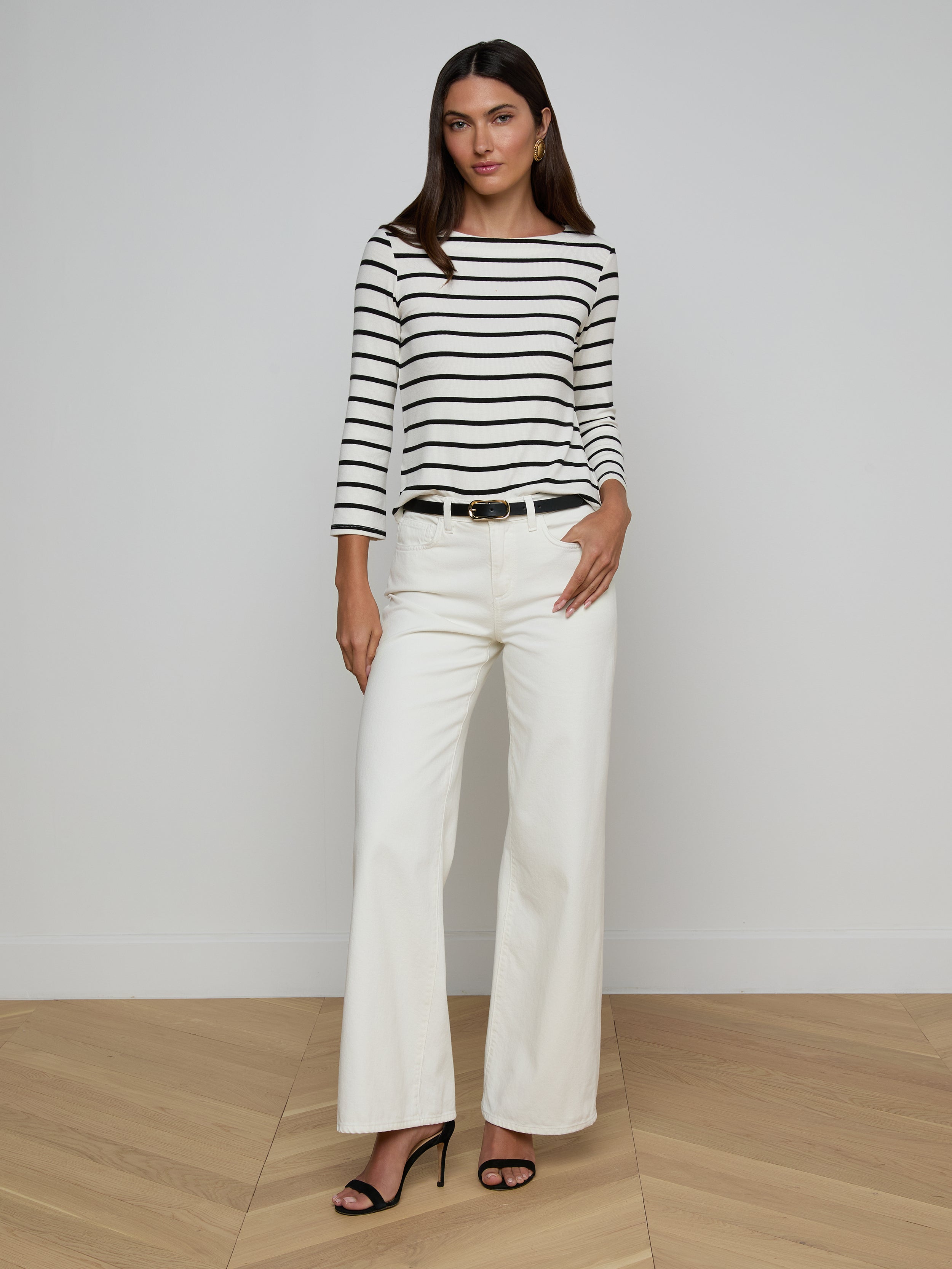 A woman stands against a plain background wearing the Lucille Striped Boatneck Top, paired with white wide-leg pants, a black belt, and black high-heeled sandals. With one hand in her pocket and a neutral expression, she captures the effortlessly chic appeal of timeless stripes.