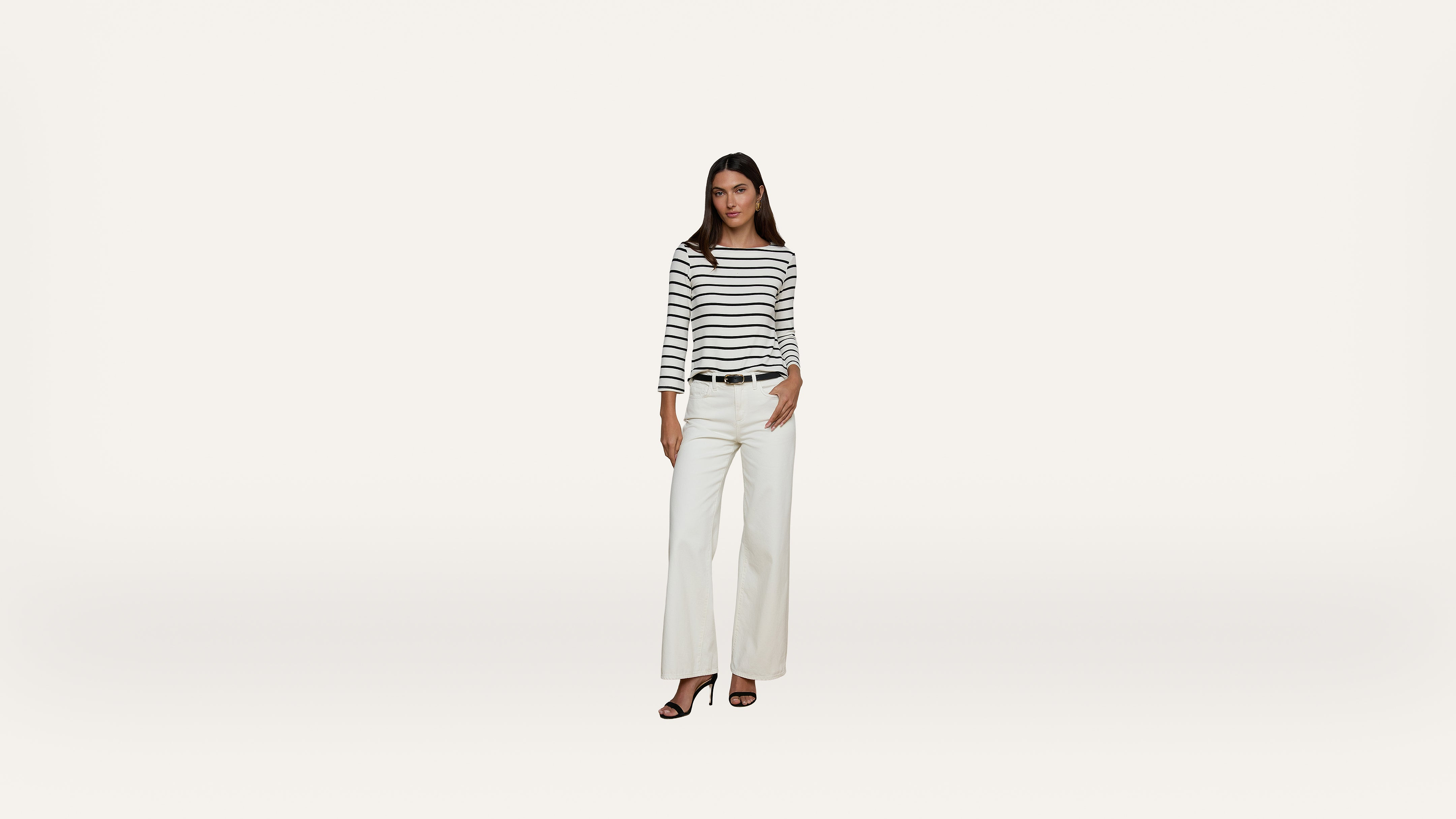 A woman wearing a black and white striped long-sleeve top, white wide-leg pants, and black sandals stands against a plain white background.