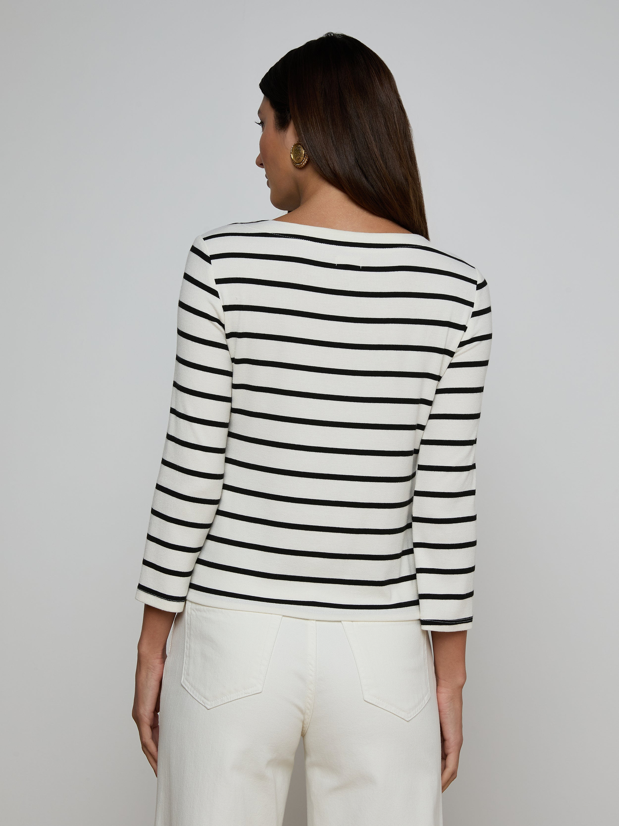 A woman faces away, wearing the Lucille Striped Boatneck Top—its stretch modal fabric features classic black and white stripes and a flattering boat neckline—paired with white pants. Her dark hair is down against a light background, with the timeless stripes completing her look.