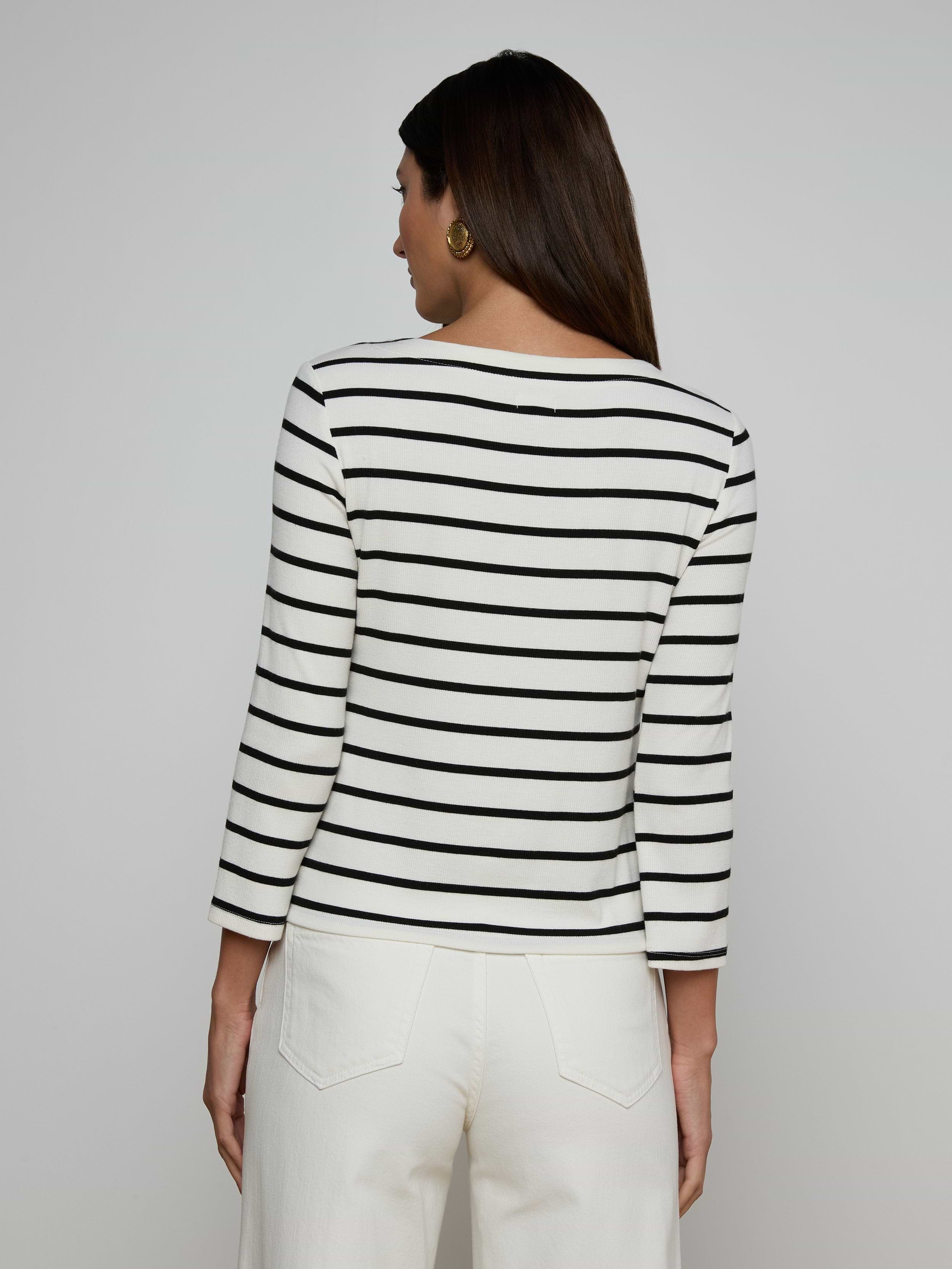 A woman faces away, wearing the Lucille Striped Boatneck Top—its stretch modal fabric features classic black and white stripes and a flattering boat neckline—paired with white pants. Her dark hair is down against a light background, with the timeless stripes completing her look.