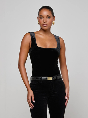 A woman poses confidently against a light background wearing the L'AGENCE Molly Velvet Bodysuit in black, styled with black pants, a gold-buckled belt, and gold hoop earrings. Her arms are relaxed as she looks at the camera.