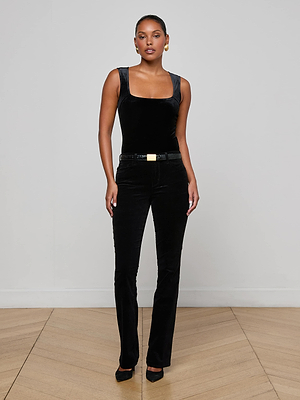 A woman stands indoors against a light wall and wooden floor, wearing the L'AGENCE Molly Velvet Bodysuit—a chic sleeveless black stretch velvet layering piece—paired with black belted pants, black heels, and gold earrings. She faces forward, hands by her sides.