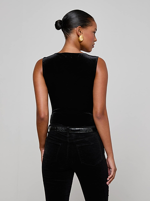 A woman with her hair in a bun and gold hoop earrings models the L'AGENCE Molly Velvet Bodysuit with black pants, facing away from the camera against a plain light gray background.