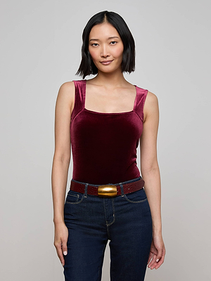 A woman with short black hair wears the L'AGENCE Molly Velvet Bodysuit in maroon with a square neckline, paired with dark blue jeans and a red belt featuring a gold buckle. She stands facing the camera against a plain light gray background.
