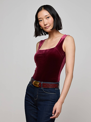 A woman with a bob haircut wears the L'AGENCE Molly Velvet Bodysuit and dark jeans with a brown belt, standing against a light gray background as she looks at the camera with a slight smile.