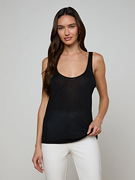 A woman with long brown hair wears the Celia Scoopneck Tank, a sleeveless black featherweight jersey top with a scoop neckline, paired with white pants. She stands against a plain light background and smiles gently at the camera.