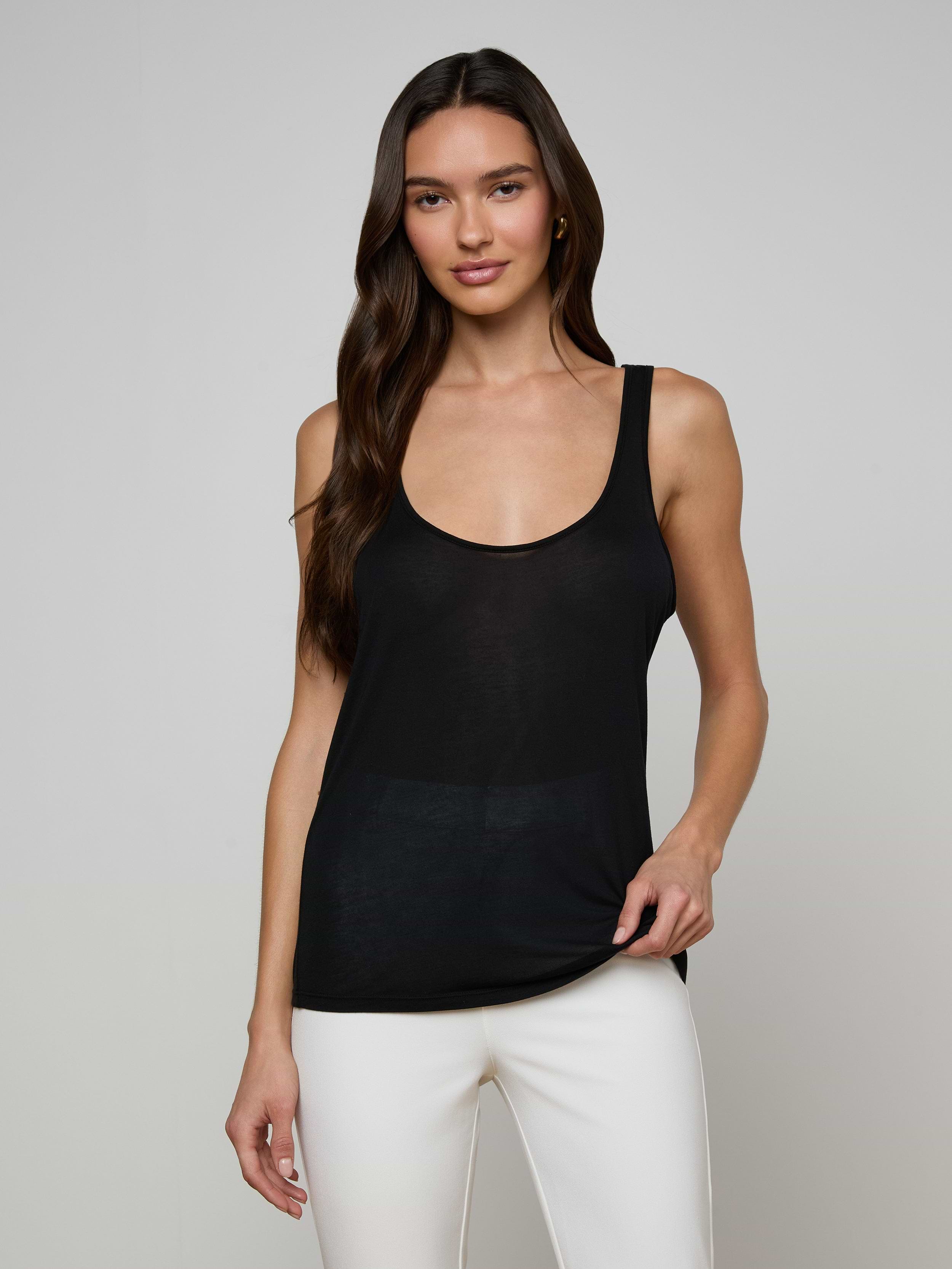 A woman with long brown hair wears the Celia Scoopneck Tank, a sleeveless black featherweight jersey top with a scoop neckline, paired with white pants. She stands against a plain light background and smiles gently at the camera.