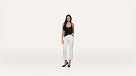 A woman stands against a plain white background, wearing a black sleeveless top, white pants, and black high heels.