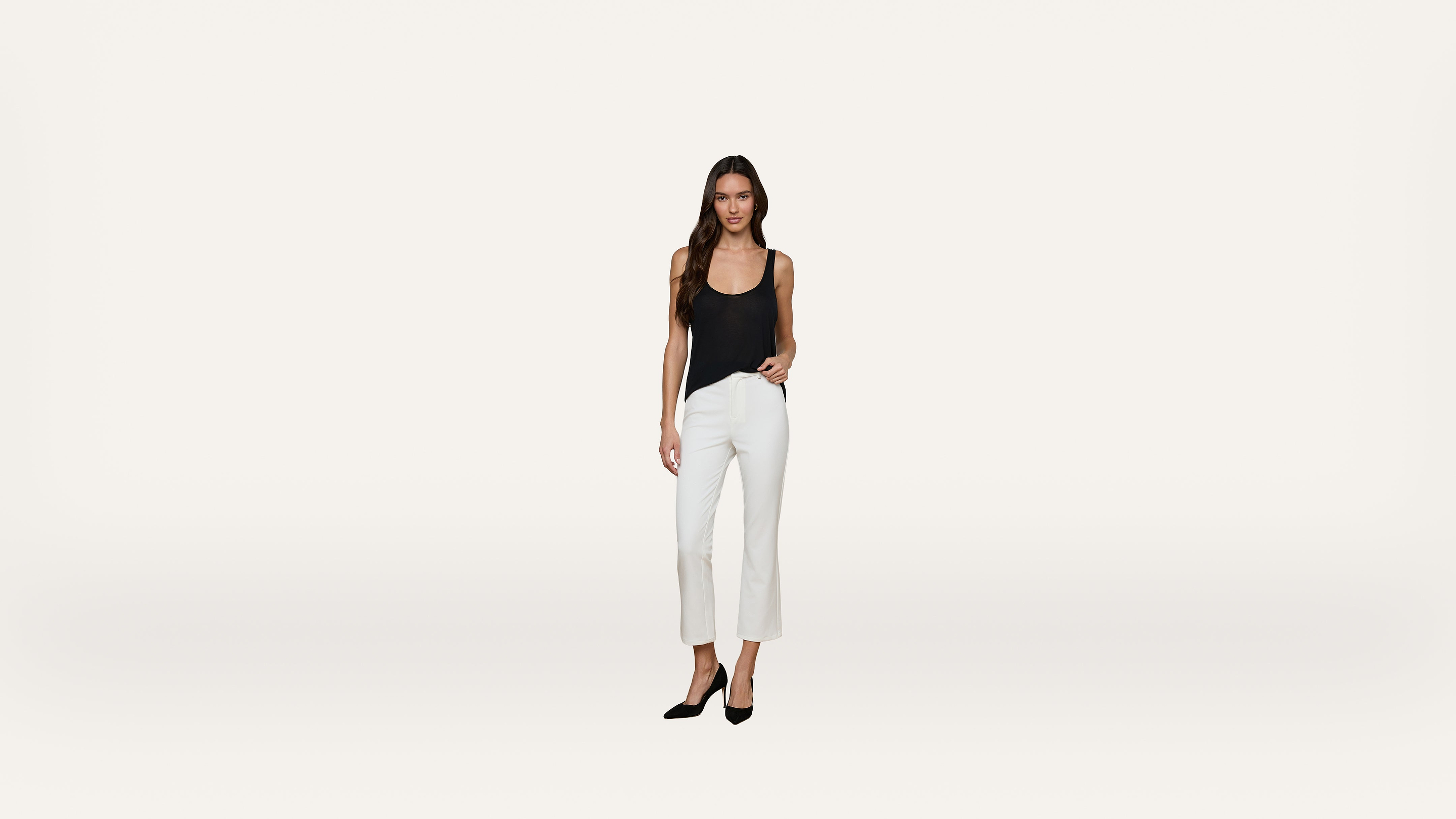 A woman stands against a plain white background, wearing a black sleeveless top, white pants, and black high heels.