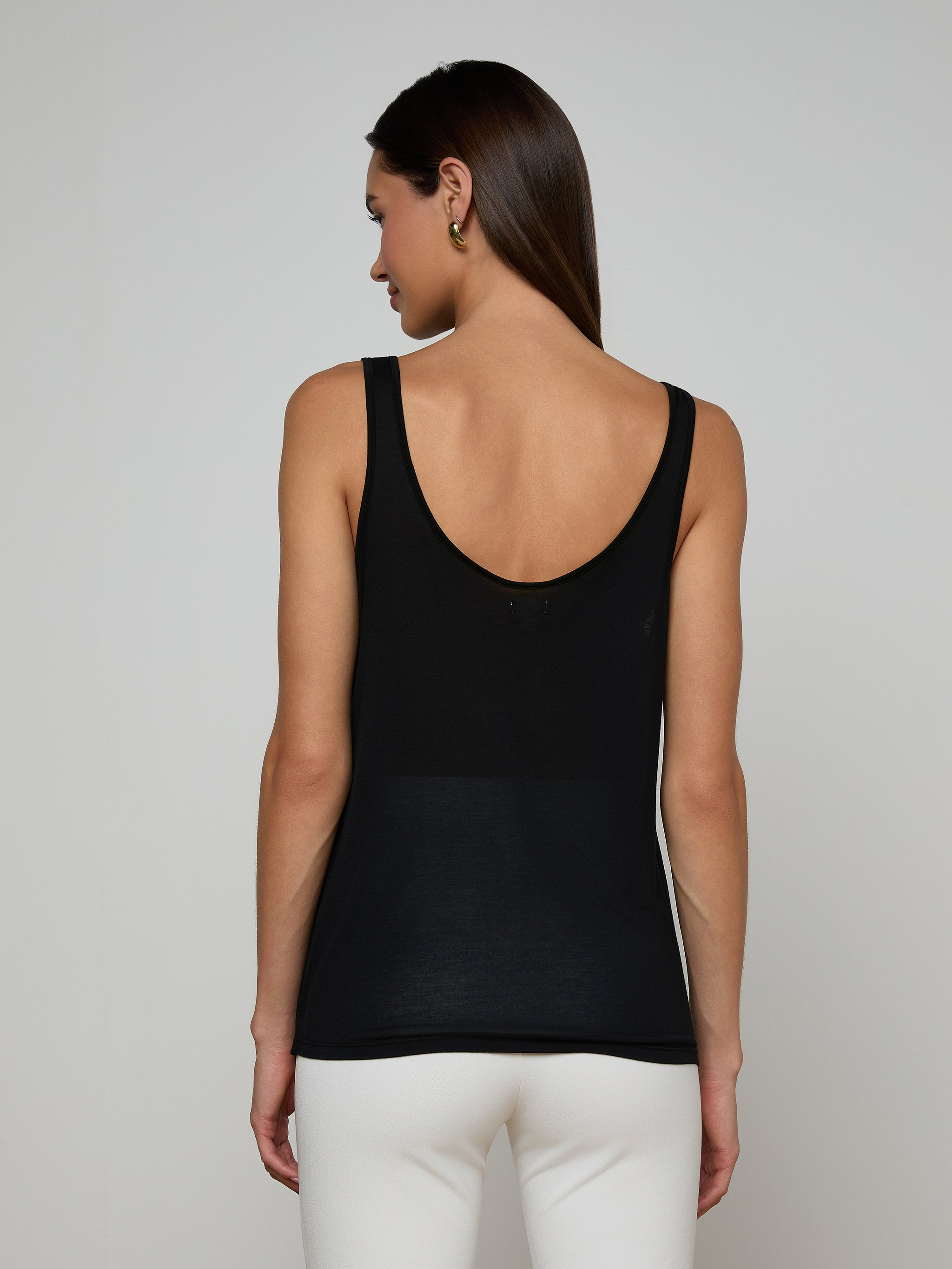 Wearing the Celia Scoopneck Tank in black, a person with straight brown hair and white pants stands with their back to the camera against a plain light background.