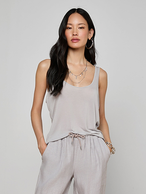 A woman with long black hair wears the Celia Scoopneck Tank in light grey with matching drawstring jersey pants. Against a plain background, she accessorizes with hoop earrings, layered necklaces, and a bracelet.