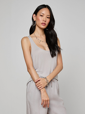 A woman with long black hair wears the Celia Scoopneck Tank in light gray, paired with matching lightweight jersey pants. She accessorizes with layered necklaces, hoop earrings, and a bracelet, standing against a plain light background.