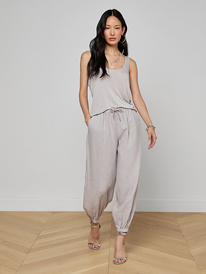 A woman stands on a light wood floor wearing the Celia Scoopneck Tank in light gray with matching drawstring pants, accessorized with layered necklaces, bracelets, and heeled sandals.