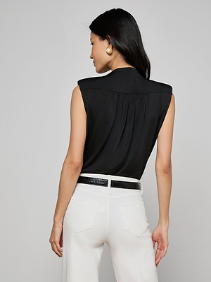 A woman with long dark hair, wearing a sleeveless black Akari Bodysuit, large round earrings, and high-waisted white pants with a black belt stands facing away from the camera against a plain light gray background.