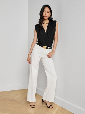 A woman with long dark hair and a neutral expression stands indoors against a white wall, wearing a sleeveless black Akari Bodysuit, white wide-leg pants, a black belt, black high-heeled sandals, and a gold necklace.