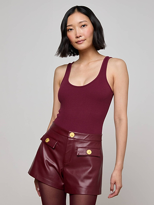 A woman with a bob haircut wears the sleeveless maroon Nica Scoopneck Bodysuit and high-waisted burgundy leather shorts with gold buttons, standing against a plain light gray background.