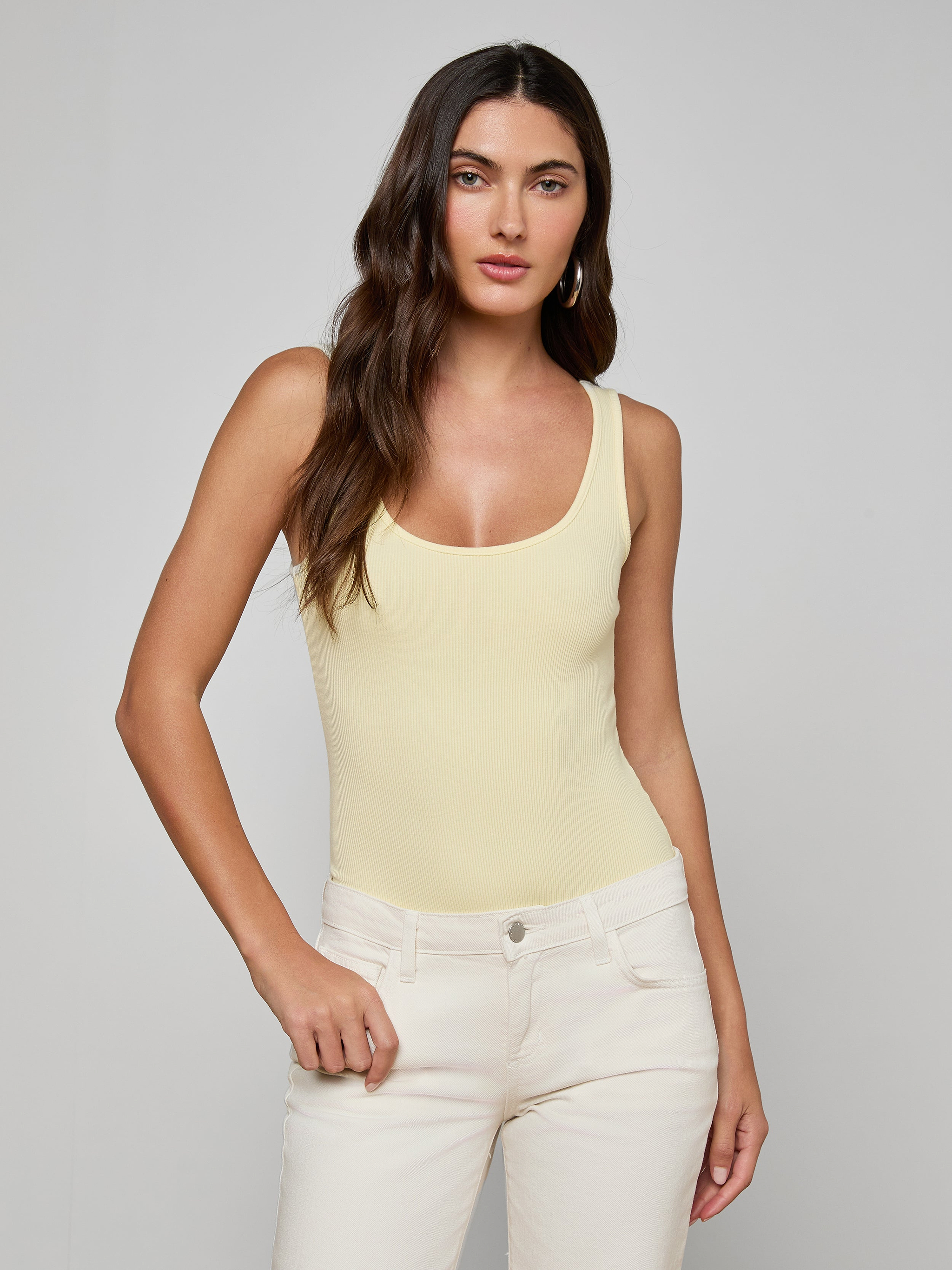 A woman with long brown hair wears a light yellow Nica Scoopneck Bodysuit with a sculpted fit and off-white pants, standing against a plain light gray background and looking at the camera neutrally.