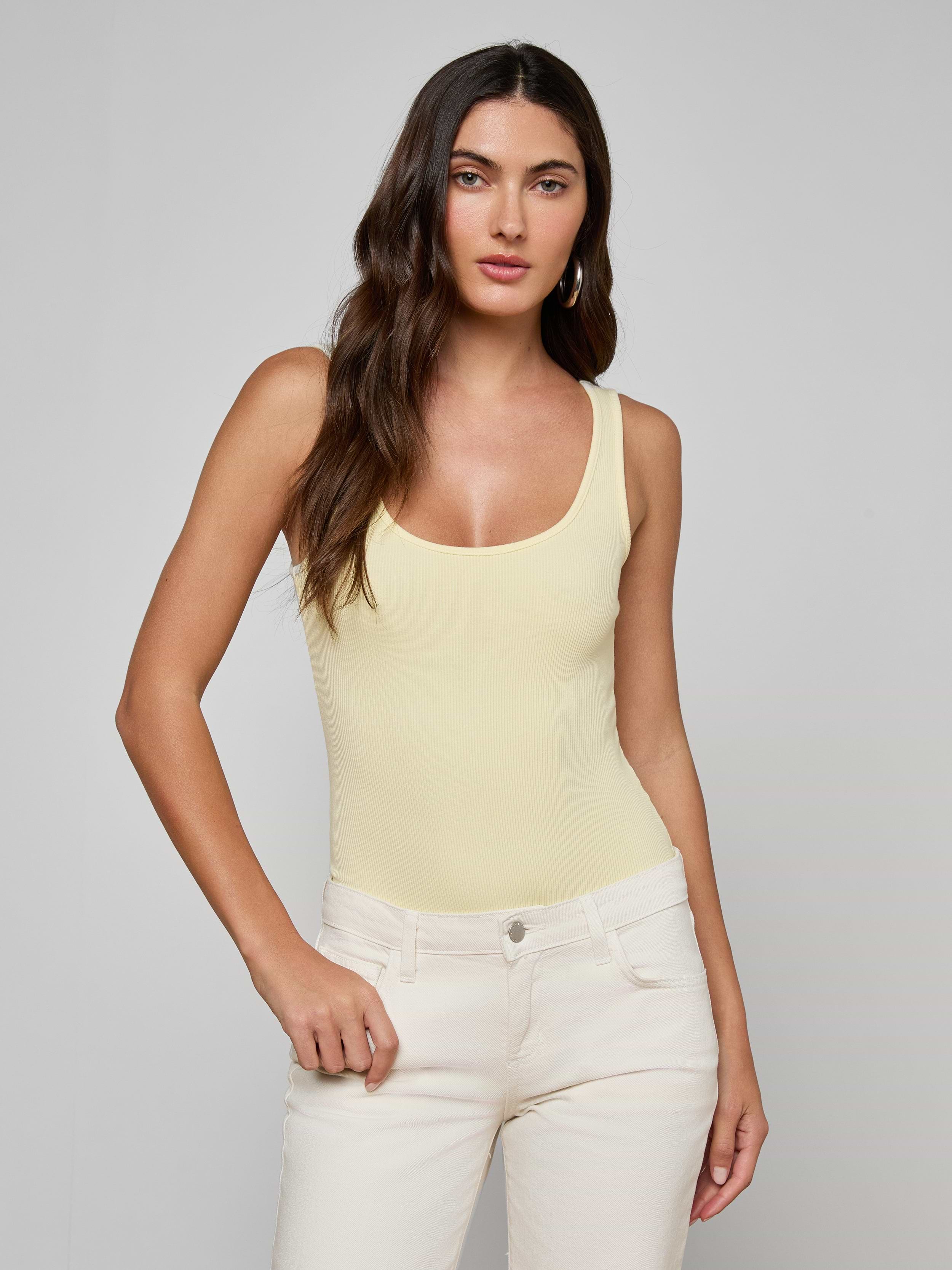 A woman with long brown hair wears a light yellow Nica Scoopneck Bodysuit with a sculpted fit and off-white pants, standing against a plain light gray background and looking at the camera neutrally.