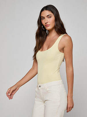 A woman with long brown hair wears a sleeveless, light yellow Nica Scoopneck Bodysuit as a base layer with high-waisted off-white pants, posing against a plain light gray background and looking at the camera neutrally.
