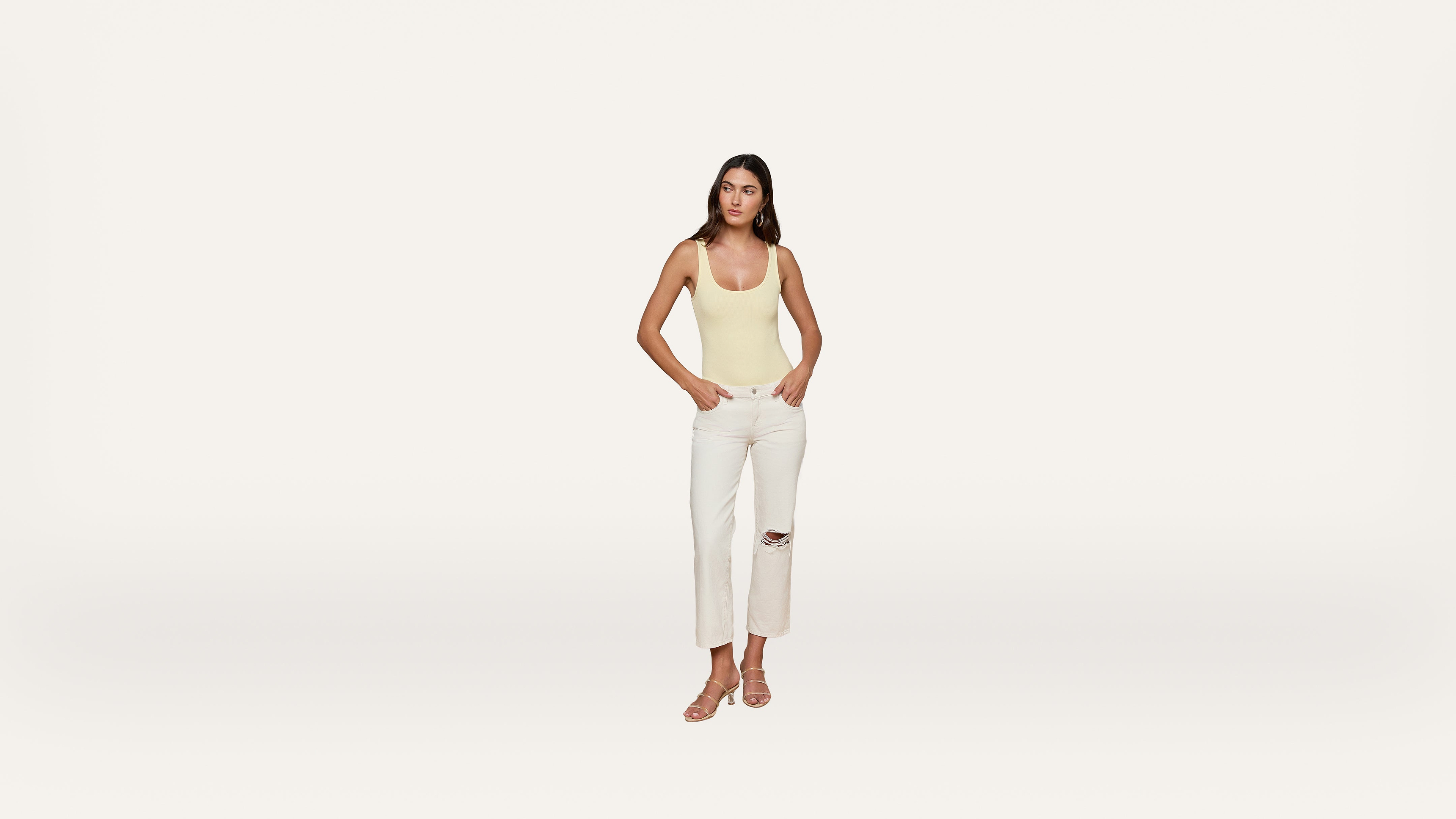A woman stands against a plain white background, wearing a yellow tank top, white ripped jeans, and light-colored sandals.