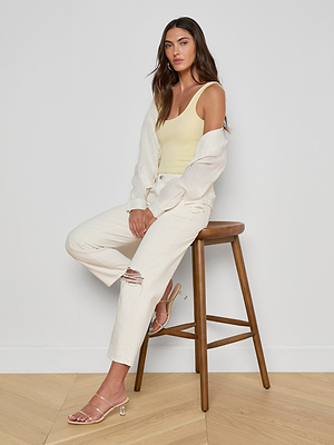 A woman with long brown hair sits on a wooden stool against a white wall, wearing the Nica Scoopneck Bodysuit in yellow under an off-white cardigan, styled with ripped white pants and clear heeled sandals for a sculpted look.