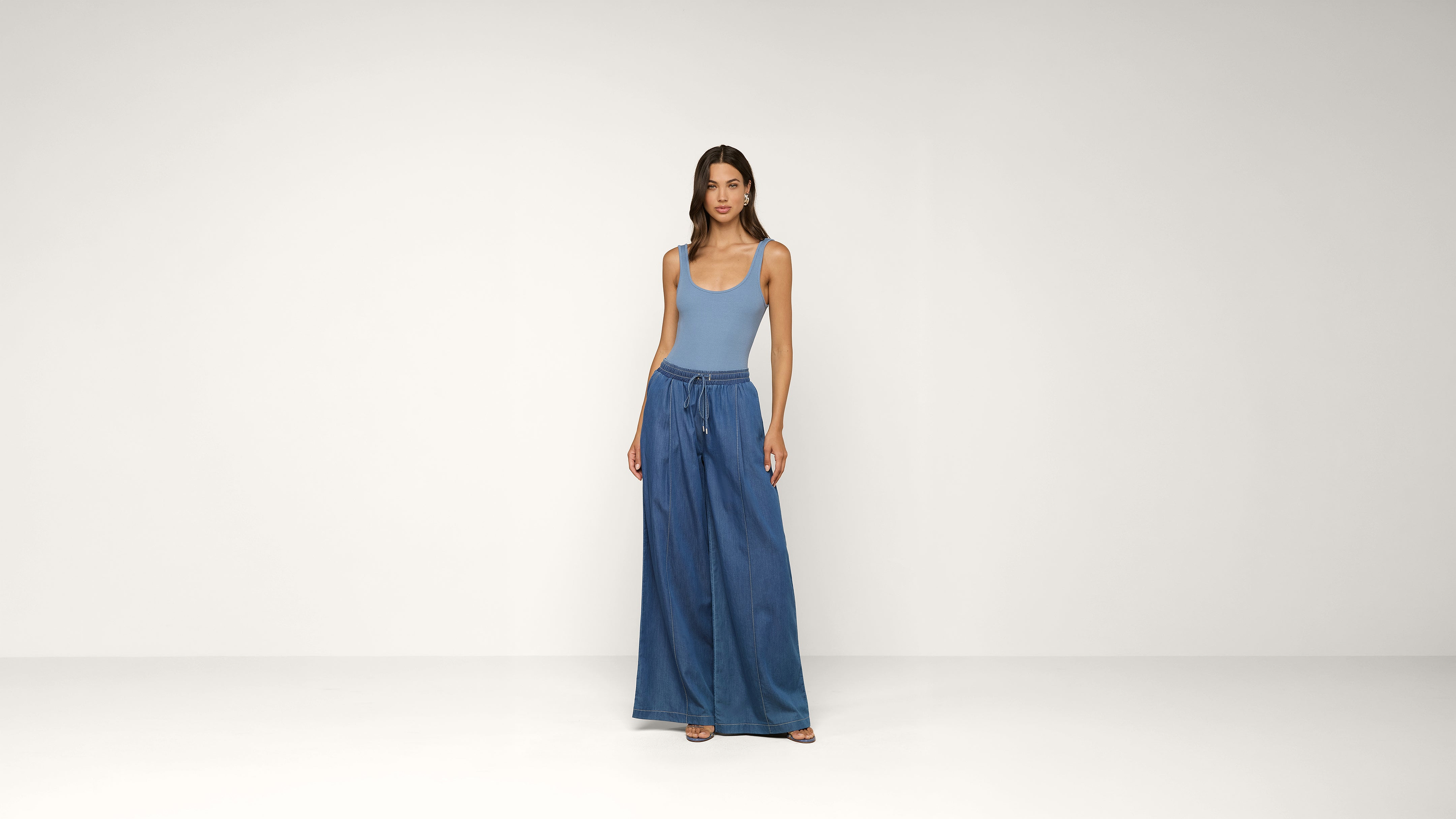 A woman stands against a plain white background wearing a light blue tank top and wide-leg blue jeans, looking at the camera with a neutral expression.