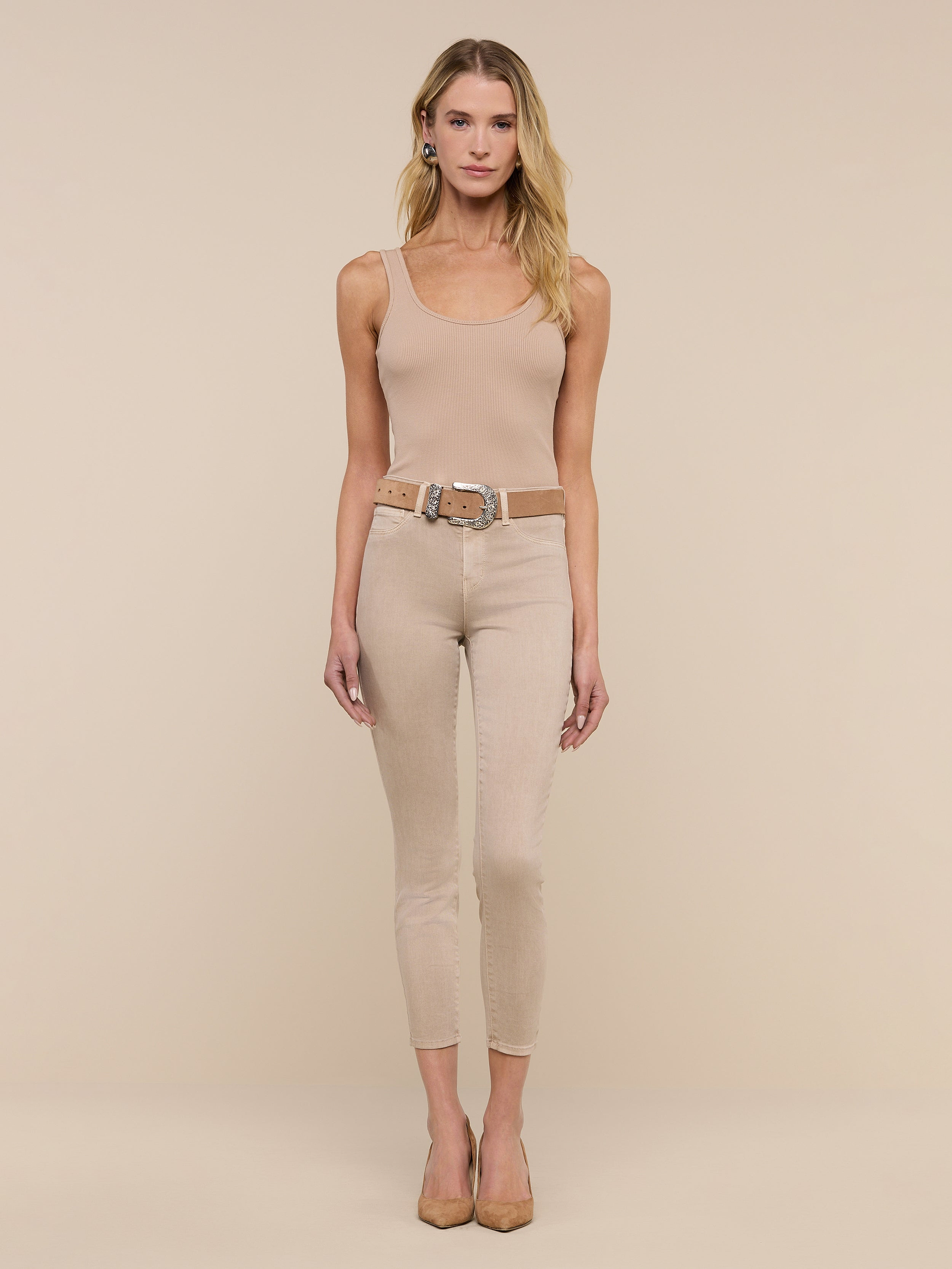 A woman with long dark hair wears the Nica Scoopneck Bodysuit, paired with light beige skinny jeans and nude heels, as she walks indoors on a light wood floor against a plain white wall.