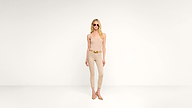 A woman wearing sunglasses, a beige tank top, beige pants, and tan heels stands against a plain white background.