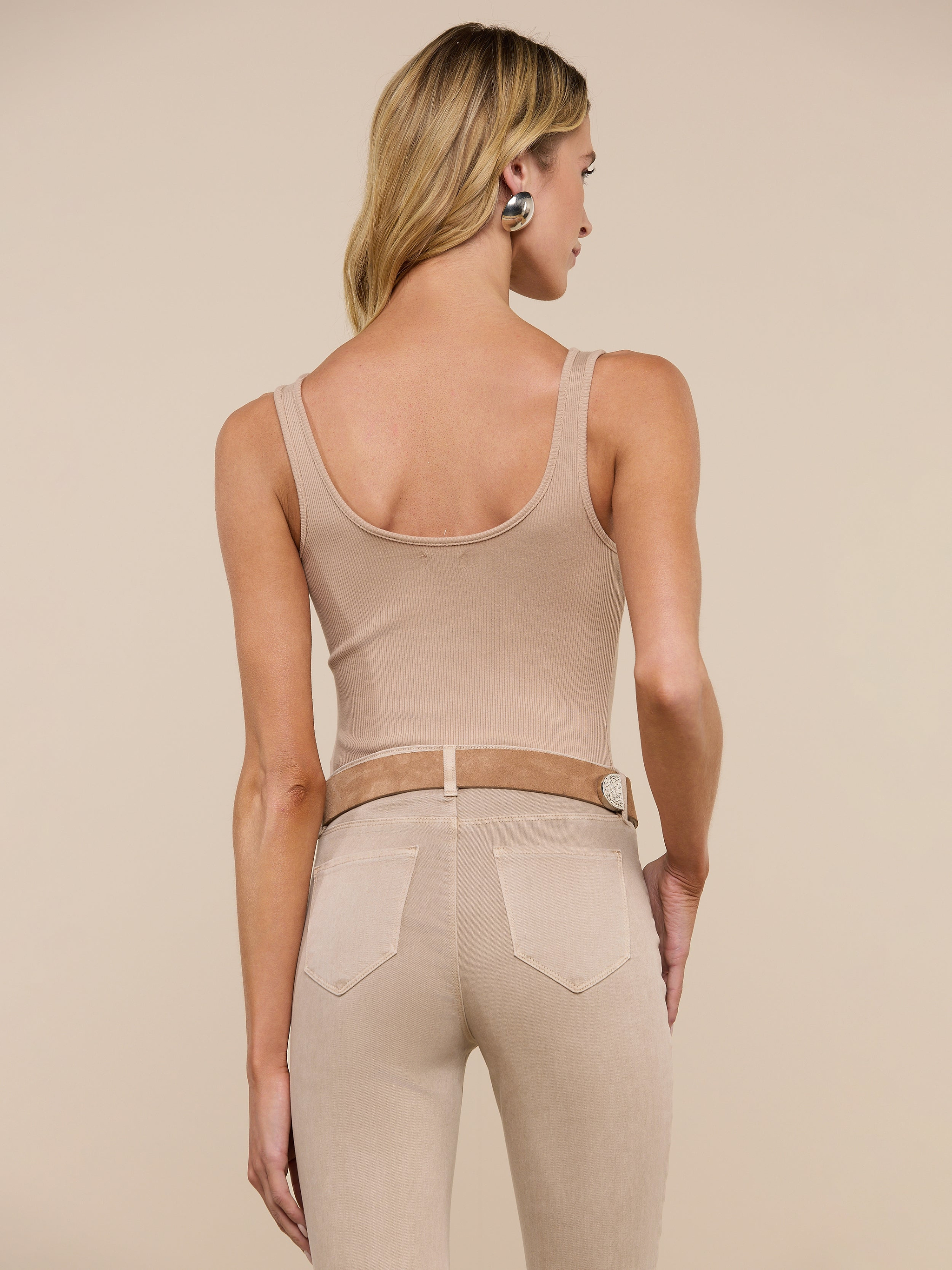 A woman with long dark hair stands with her back to the camera, wearing a beige Nica Scoopneck Bodysuit and light tan jeans against a plain light gray background.