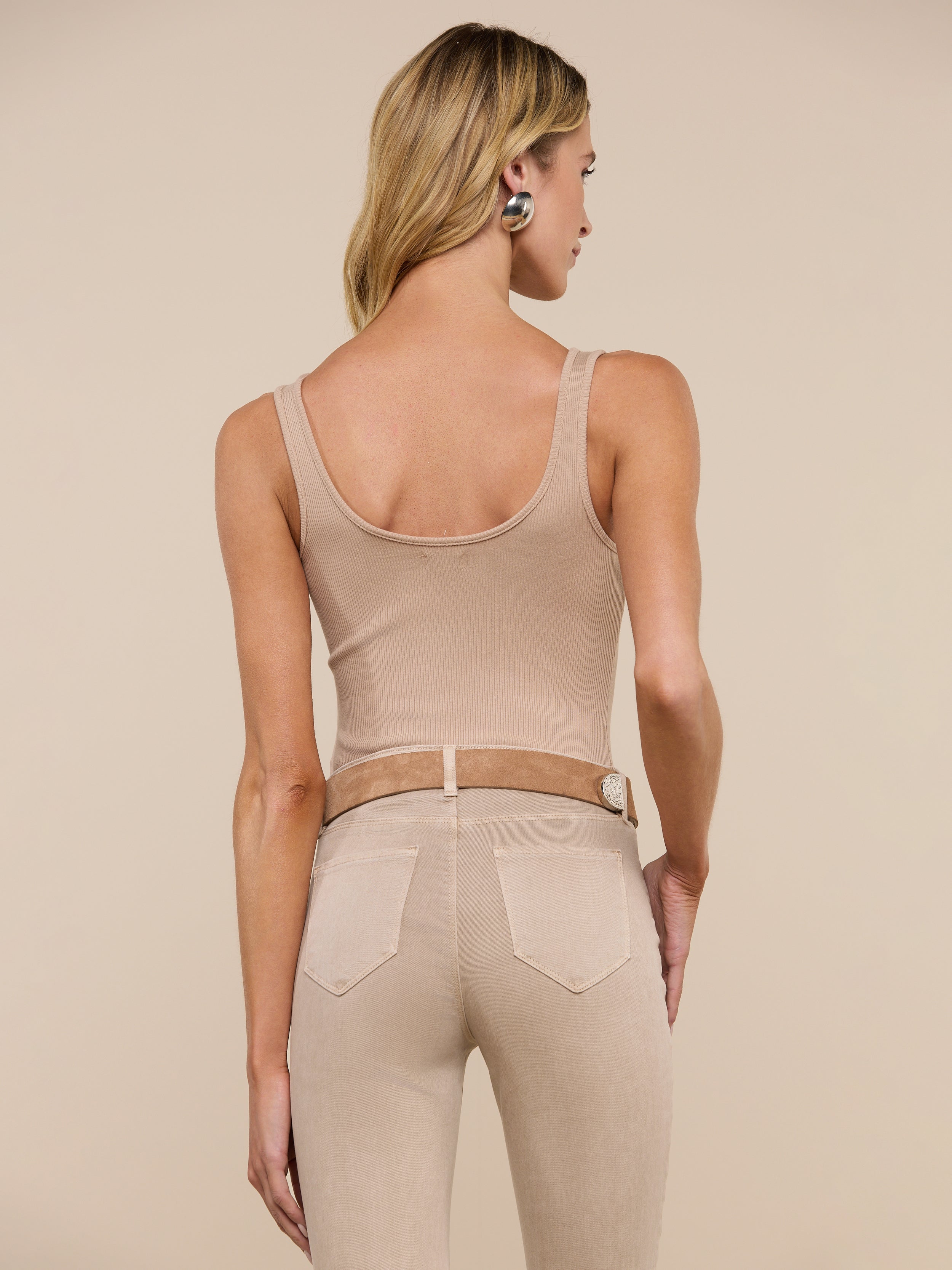 A woman with long dark hair stands with her back to the camera, wearing a beige Nica Scoopneck Bodysuit and light tan jeans against a plain light gray background.