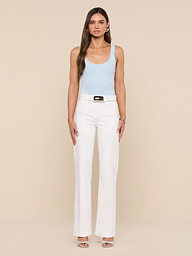A woman stands facing forward against a plain white background, wearing a light blue Nica Scoopneck Bodysuit with a sculpted fit, paired with white high-waisted pants featuring a belt and open-toed heels.