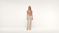 Woman in beige tank top and wide-leg pants stands facing forward in a minimalist, white studio setting. She wears sunglasses, a gold belt, and heeled shoes. The background is plain and unadorned.