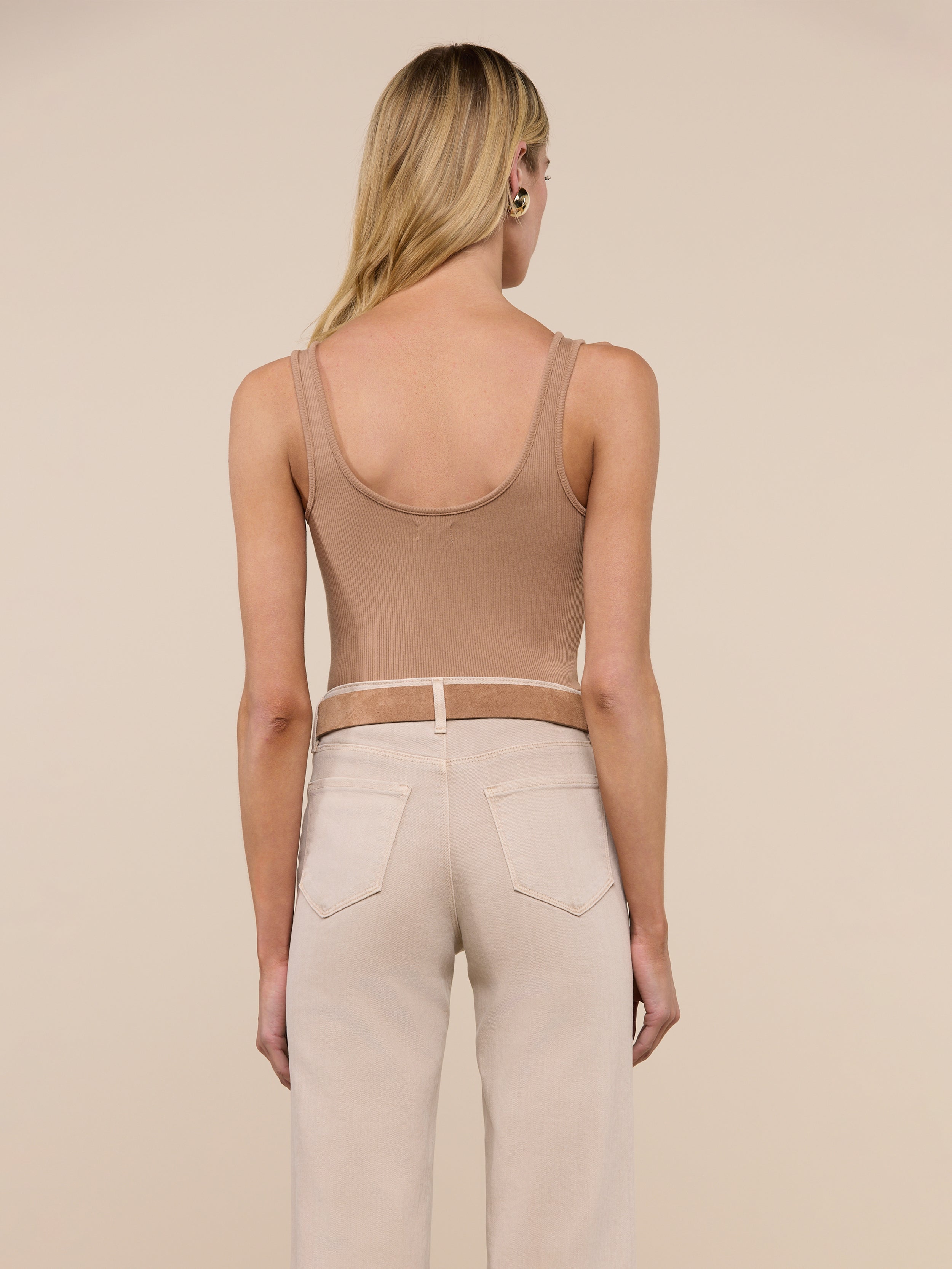 A woman with long blonde hair, dressed in a tan Nica Scoopneck Bodysuit as a stretch rib base layer and light beige pants, stands facing away from the camera against a plain light background.