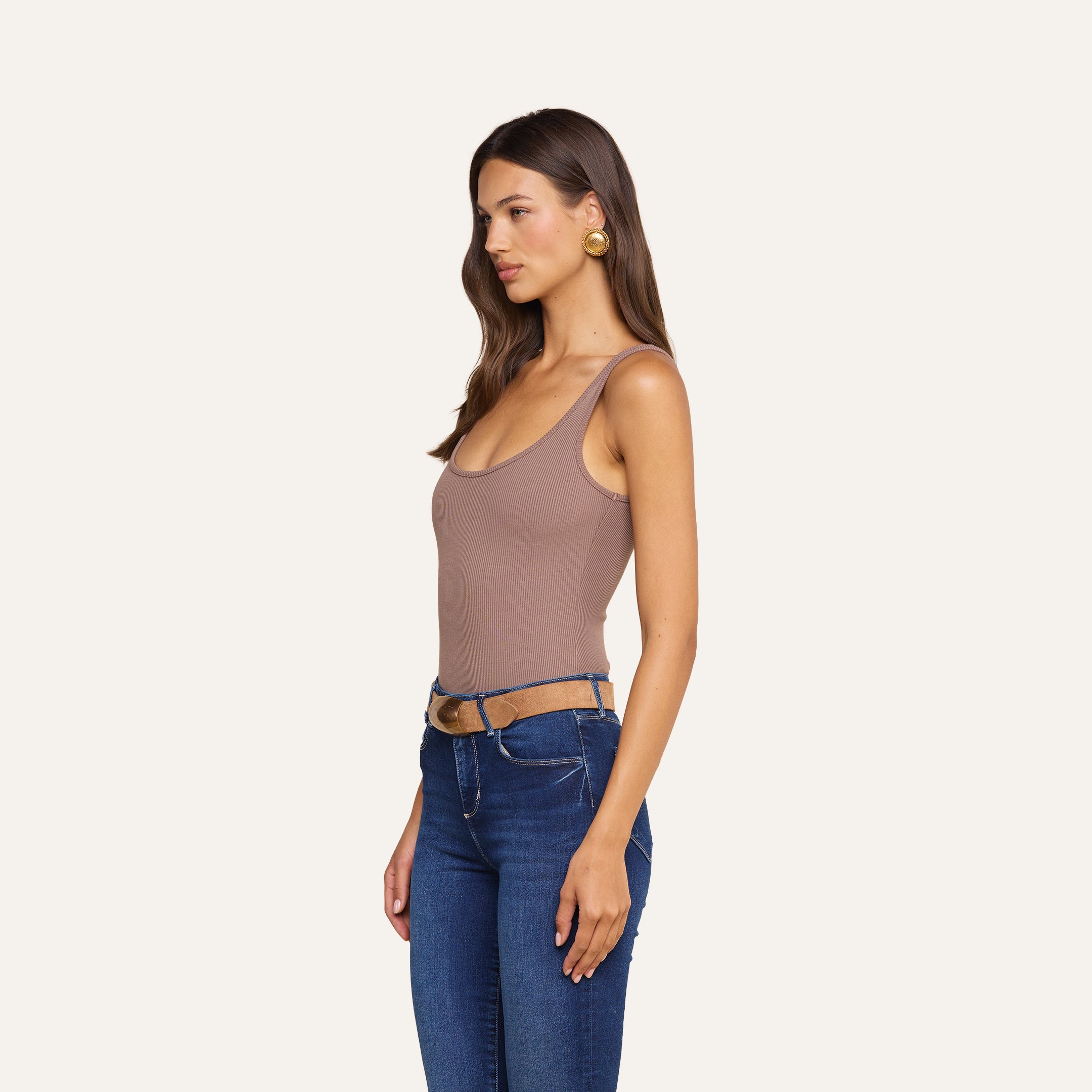 Une femme aux longs cheveux châtains, portant des boucles doreilles en or, est vêtue dun body Nica à encolure dégagée de couleur taupe, dun jean bleu et dune ceinture beige clair. Elle se tient debout, détendue, tournée vers la gauche, devant un fond beige uni.
