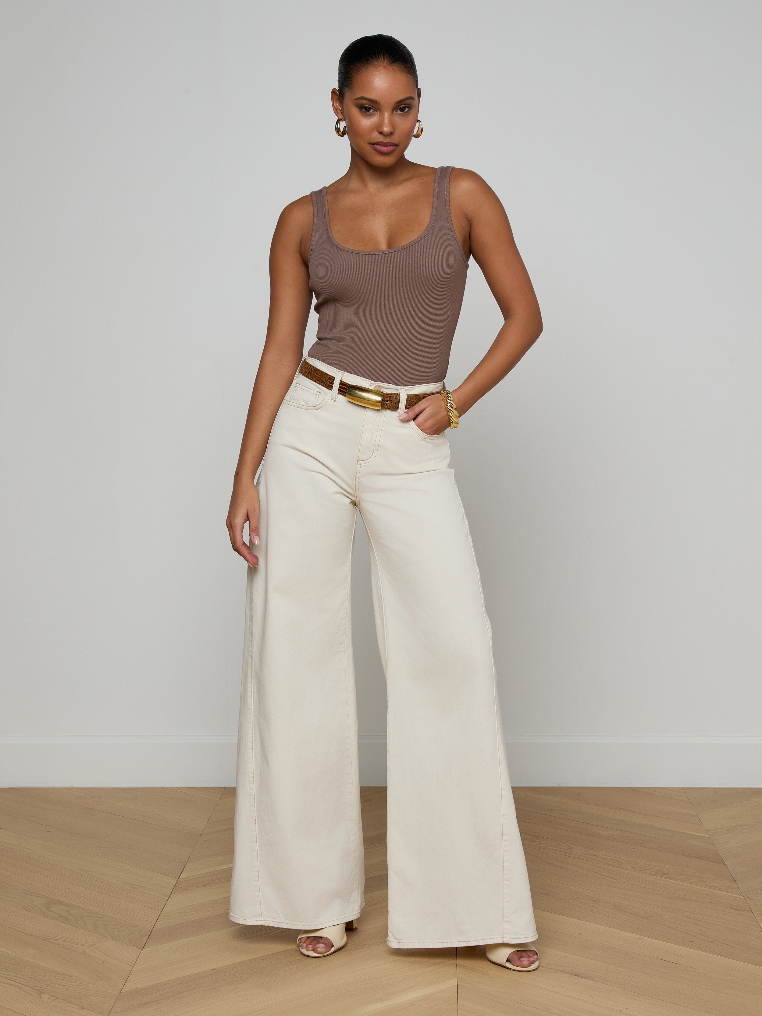 A woman wearing sunglasses, the Nica Scoopneck Bodysuit in taupe with a stretch rib and sculpted fit, blue cropped jeans, and nude heels stands against a plain white background.