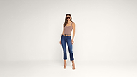 Woman wearing sunglasses, a sleeveless mauve bodysuit, blue cropped jeans with a belt, and nude heels stands against a plain white background.