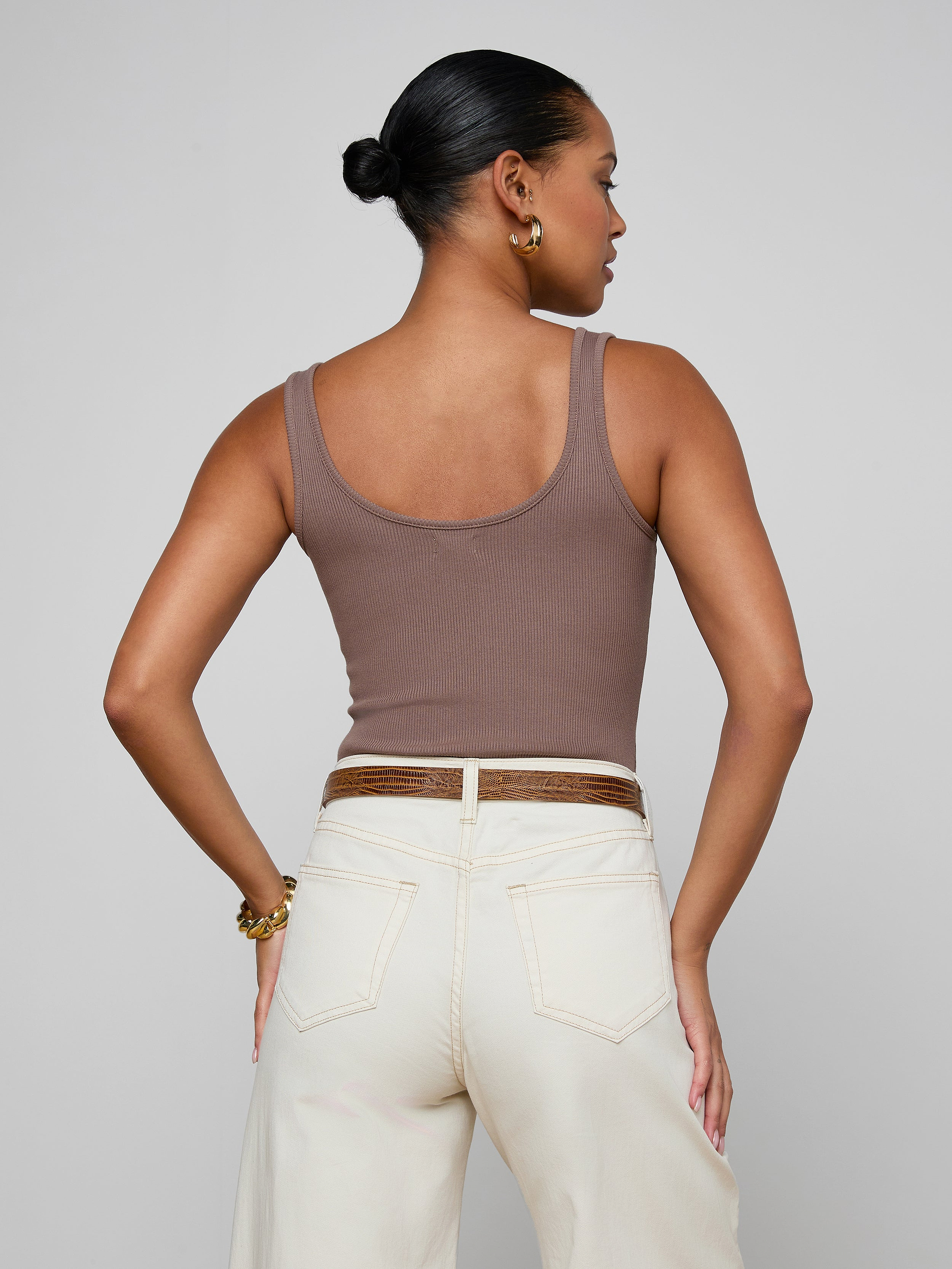 A woman with long brown hair is shown from the back, wearing the Nica Scoopneck Bodysuit in a sculpted, sleeveless fit paired with high-waisted blue jeans featuring a visible waistband. The background is plain and light-colored.