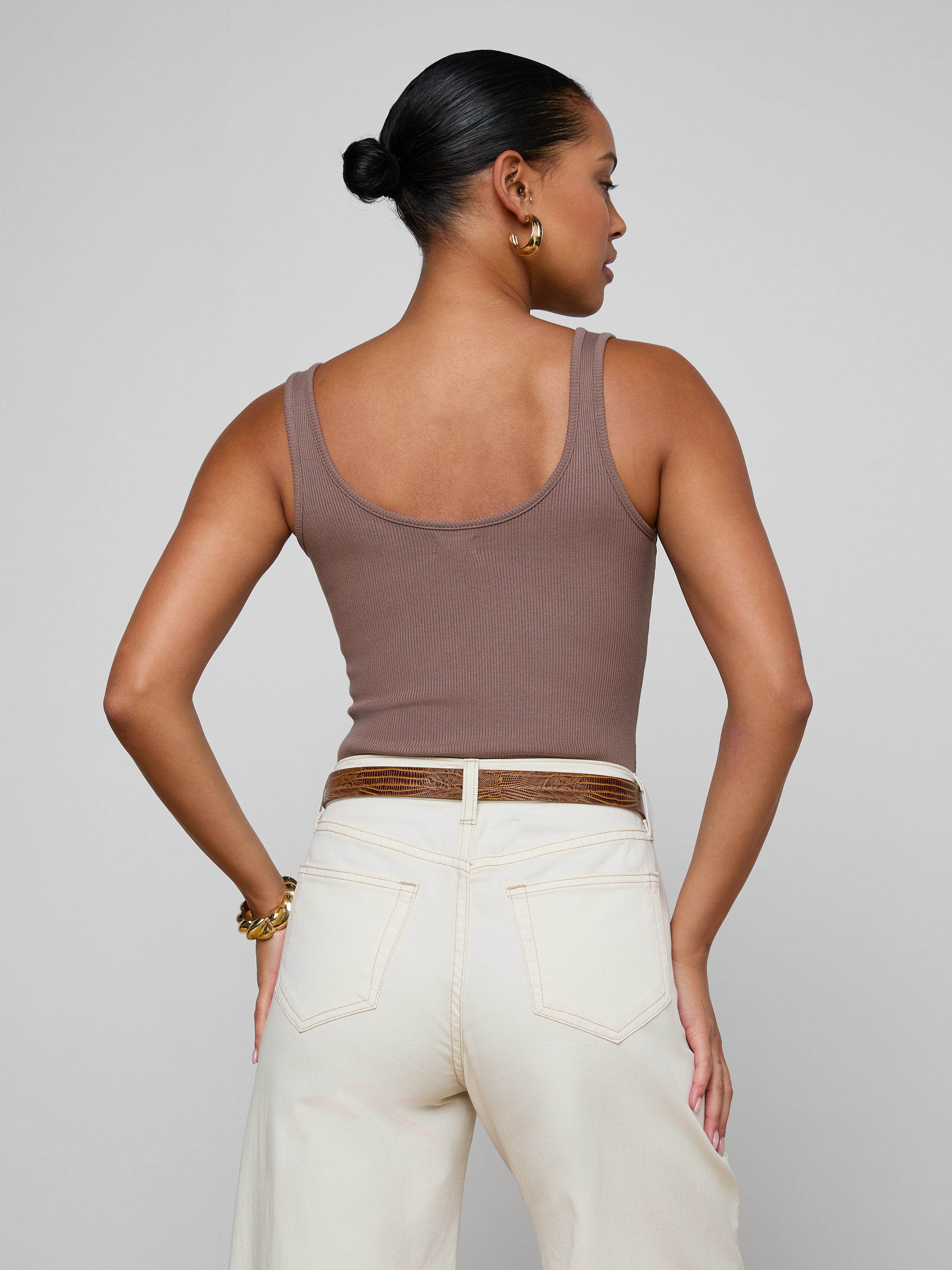 A woman with long brown hair is shown from the back, wearing the Nica Scoopneck Bodysuit in a sculpted, sleeveless fit paired with high-waisted blue jeans featuring a visible waistband. The background is plain and light-colored.