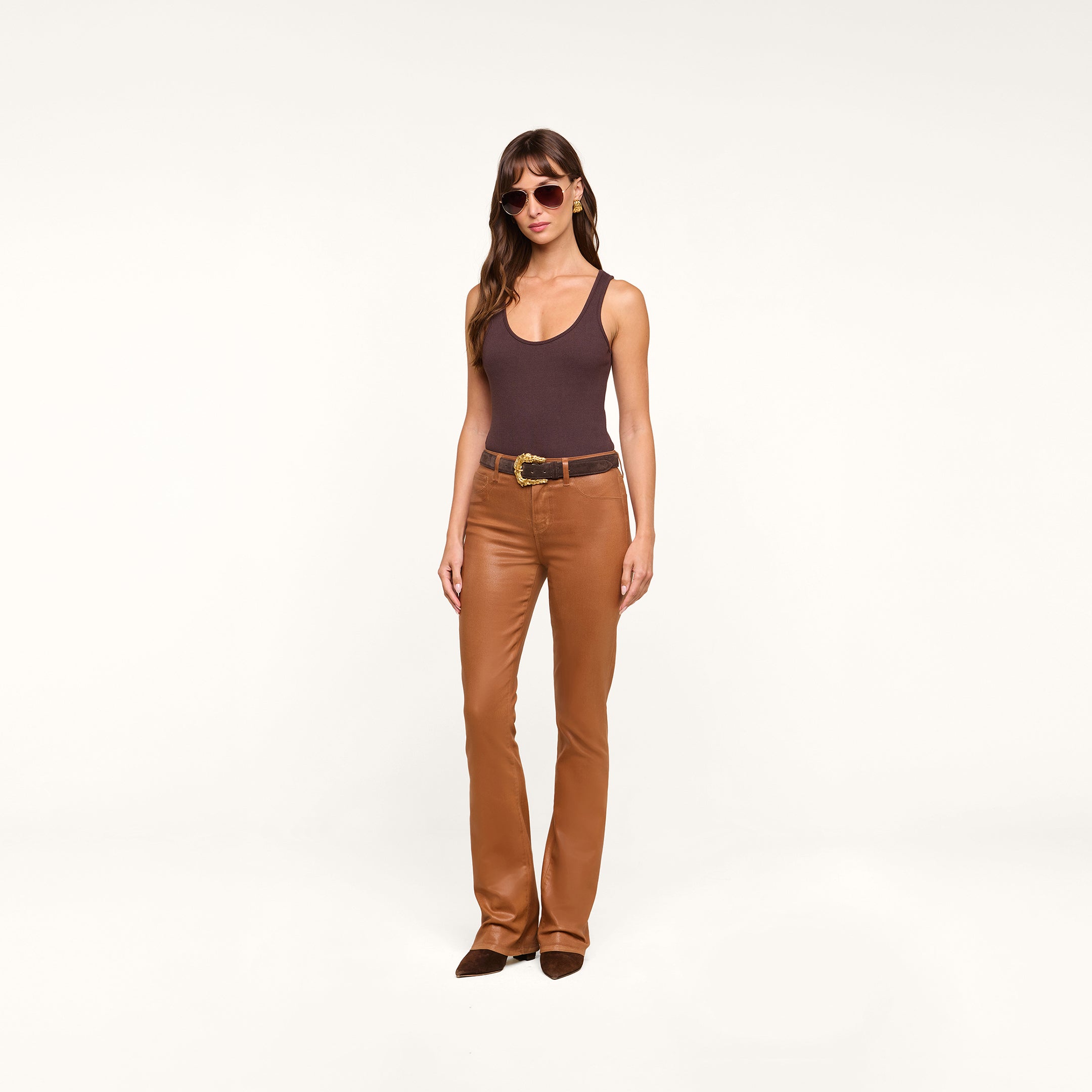 A woman wearing sunglasses, a dark brown Nica Scoopneck Bodysuit, brown leather pants with a belt, and brown shoes stands against a plain white background.