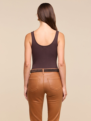 A woman with long brown hair, wearing a sleeveless dark brown Nica Scoopneck Bodysuit that offers a sculpted fit and high-waisted tan leather pants, is standing with her back to the camera against a beige background.
