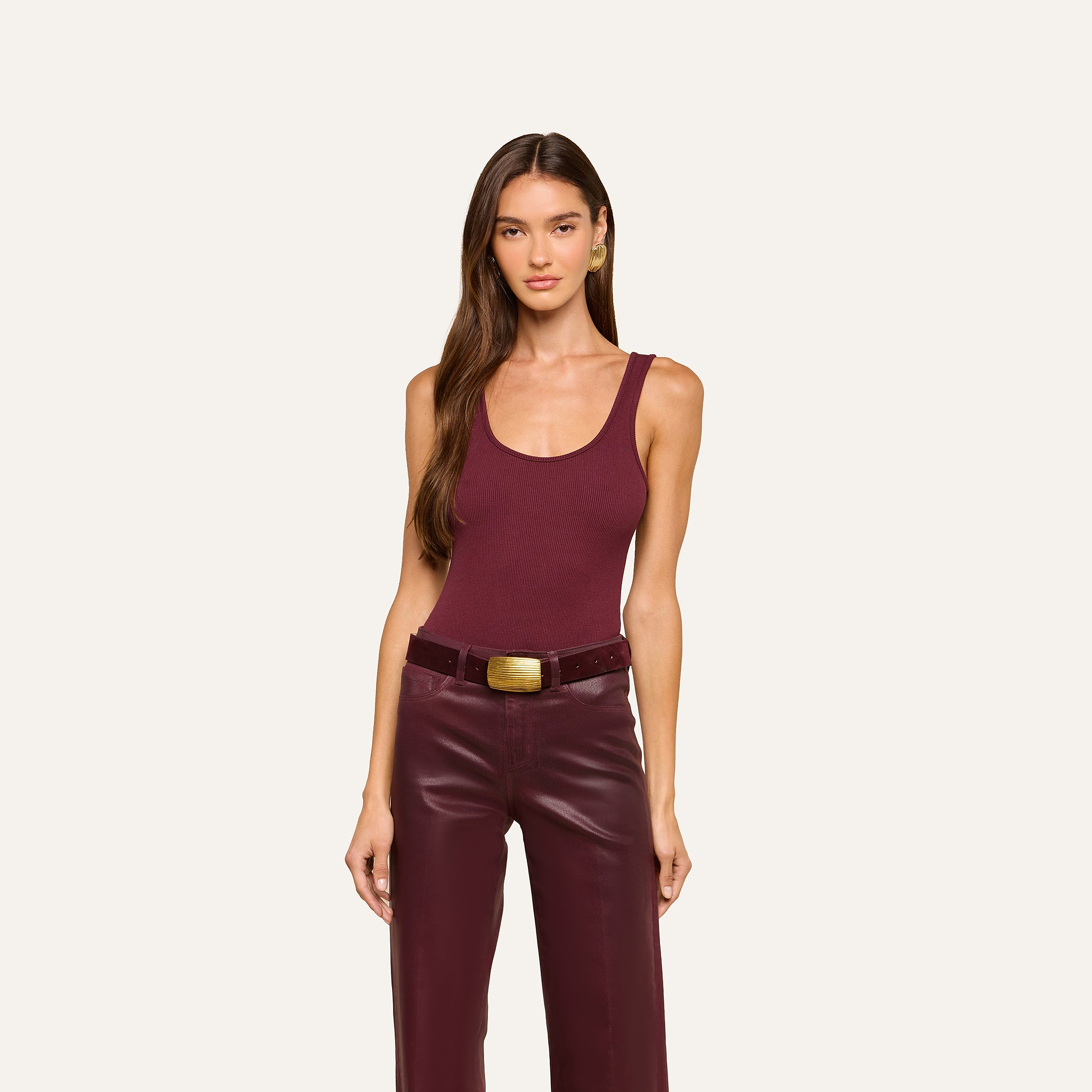 A woman with long brown hair wears a sleeveless maroon Nica Scoopneck Bodysuit paired with matching leather pants and a wide belt, standing against a plain light background, looking forward with her arms relaxed at her sides.