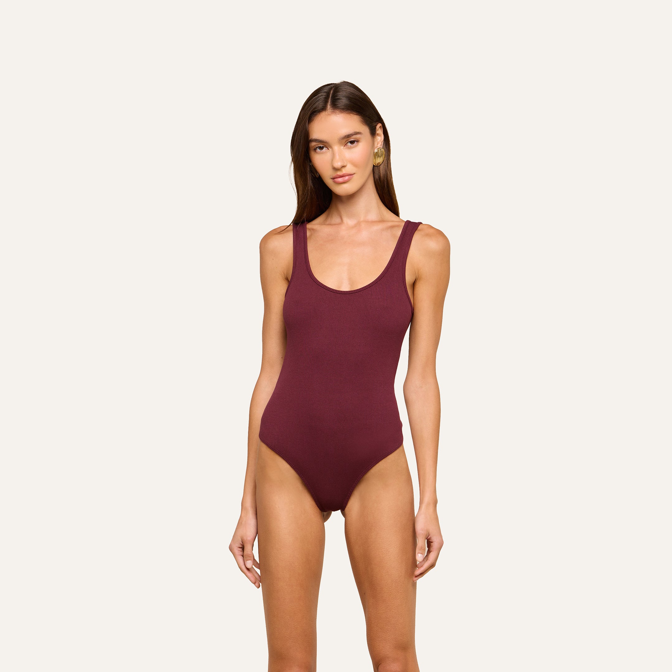 A woman faces forward against a plain light background, wearing the sleeveless Nica Scoopneck Bodysuit in dark maroon with high recovery stretch.